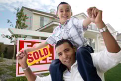 Hispanic Father and Son in Front of Their New Home with Sold Home For Sale Real Estate Sign.
