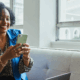 Smiling real estate agent sitting on a couch, looking at her smartphone with a laptop nearby, preparing marketing video content.