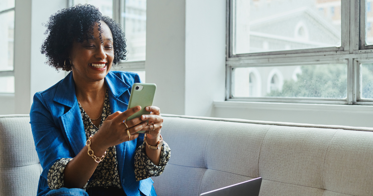 Smiling real estate agent sitting on a couch, looking at her smartphone with a laptop nearby, preparing marketing video content.