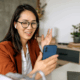Smiling real estate agent in glasses and blazer holding phone on a video call, sitting at a desk with laptop and papers, building trust with clients online.